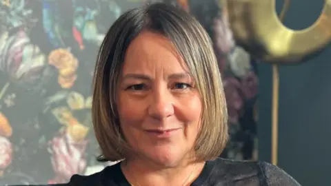 A head shot of Sue Greenwood. She has mid-length blonde/brown hair and is sitting on a sofa in front of green floral wallpaper. She is wearing a long sleeve black top and a gold necklace. 