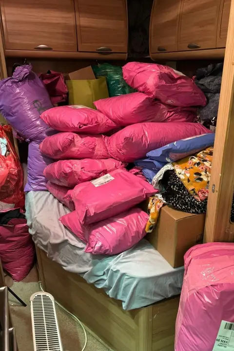 Rachel Grady Stacks of pink parcels can be seen piled on top of a double bed within a caravan. The parcels pile up to the bottom of overheard cupboards, which are a dark wood colour. 