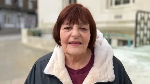 Sam Read/BBC Hazel Simmons in a dark jacket with a fleece collar standing outside, near a light-coloured building with columns and railings.