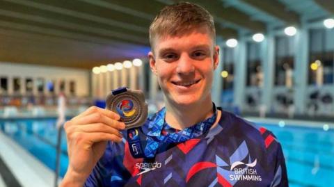 Swimmer Olly Morgan smiles whilst wearing a British Swimming branded top and holding a medal up for the camera