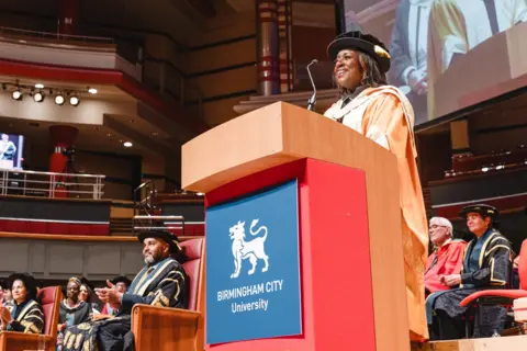 BCU Marverine Cole stands behind a lectern which has a blue sign saying Birmingham City University on it, with the crest of a lion. She wears a yellow and white graduation gown and black cap. She smiles as she talks into a microphone while on stage. Several other people in gowns are seated behind her.