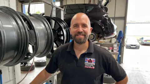 Alan Piggott smiles at the camera as he is photographed in his garage. A black car is risen on a platform behind him and he is standing next to a row of hanging alloy wheels. He is wearing a black polo neck and has a beard.  