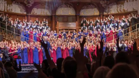 Choir performers are lit up on stage in front of an organ, wearing red and blue concert outfits.