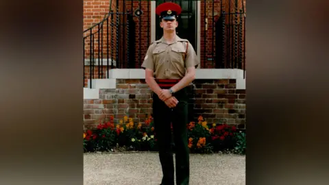 Combat Stress An older photo of a young man standing up tall, with a light brown top on, black trousers and a red and black belt, with a red and black military hat on.
