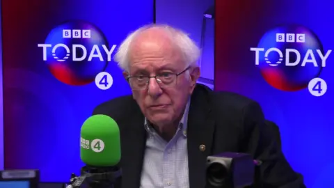 Bernie Sanders is wearing a dark suit. He is sat behind a green Radio 4 branded microphone and in front of a blue wall.