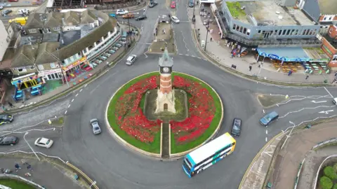 An angled birdseye photo of a clock tower. It acts as a roundabout and is surrounded by green grass which is covered in poppies. There are multiple cars and a bus going round the roundabout.