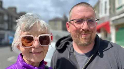 On the left is an older woman with short grey hair and pink framed sunglasses on. She's smiling and wearing a purple coat. Next to her is a man with short grey hair and a grey and ginger beard. He's wearing glasses, a grey t-shirt and grey hoodie. 