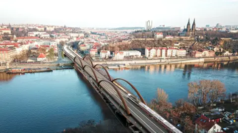 Sprava zeleznic ('Railway Authority) An architectural visualisation showing the proposed replacement bridge spanning the River Vltava in Prague . Like the original it is a bronze colour and has three large arches on either side of the bridge.