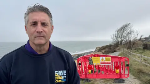 Andrew Turner/BBC A man with grey hair wearing a navy sweatshirt with Save Hemsby Coastline on the left side of his chest. He is standing on a cliff top, with a grey sea beyond. Behind him is red plastic hoarding arranged in a triangle shape with a yellow sign on it saying "caution coastal erosion"