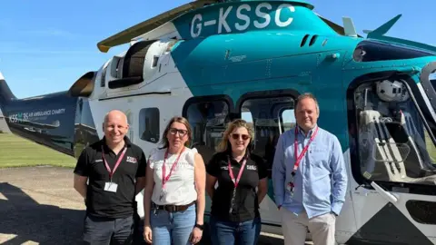 Duncan Bootland, Sophy Dunn, Helene Poursain and Warren Porritt smiling for the camera in the sun. They are stood beside a blue and white KSS helicopter.