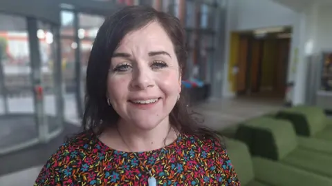 Carina is looking into camera and has a big smile. She is wearing a black dress with small flowers that are blue, pink and orange. She has long black hair which is tied back and she wears a crystal pendant. She's standing in the lobby of Cardiff University's CUBRIC building where the study is taking place. 