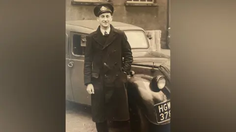 Robin Page A black and white image of a postman in the post-war era in the late 1940s. He wears a long black coat, black hat, tie, white shirt and is standing in front of a car. 