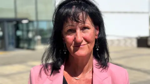 Jackie Hallam, who has shoulder-length black hair and wears a pink jacket over a black blouse, is pictured outside the courthouse in the Isle of Man.