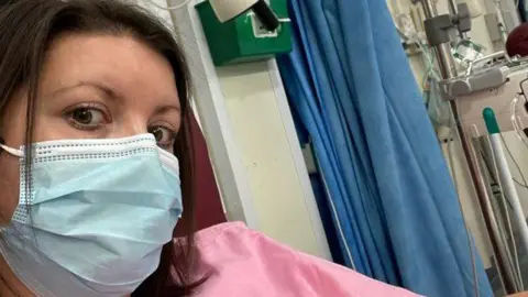 Handout Carly looks into the camera with a blue mask over her nose and mouth. She is sitting in a hospital bay. 