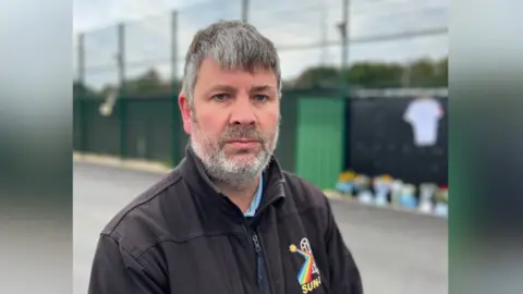 George Carden/BBC James Wilson posa una foto en el estadio de fútbol Chichester FC. James tiene cabello negro/gris y usa una chaqueta de cremallera negra y una camisa azul. El tributo floral y de la tarjeta se puede ver en el fondo, aunque parcialmente borrosa.