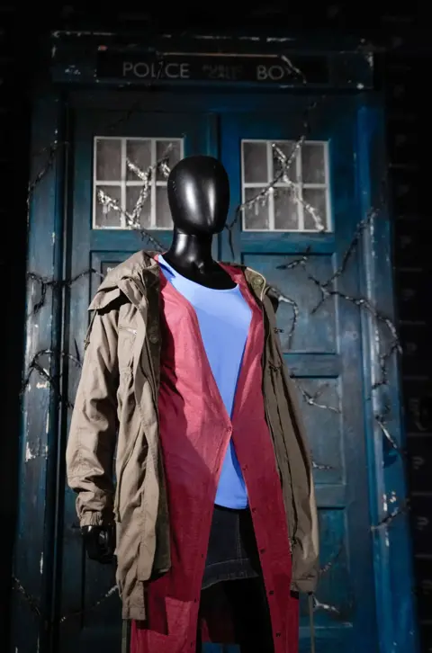 PA Media An Amy Pond costume from Doctor Who series five, episode seven: Amy's Choice. Displayed on a mannequin, the costume comprises a denim skirt, a white t-shirt, a long red cardigan, and a brown parka