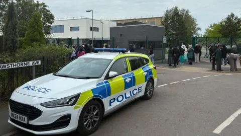 BBC A police car is parked up outside Cedar Mount Academy as well as a number of people waiting outside the entrance.