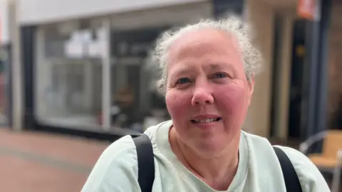 A woman with tied-up gray hair wearing light green sweatshirts and black backpacks and smiling on camera, in front of the shops behind HR.