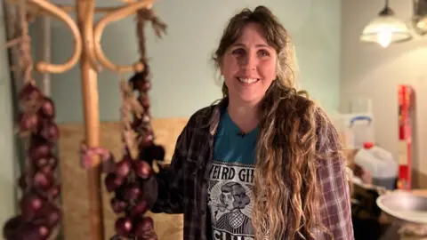 Josie wears a red checked shirt over a blue t-shirt which says 'Weird Girls', smiling toward the camera with a hatstand holding rows of onions behind her. 