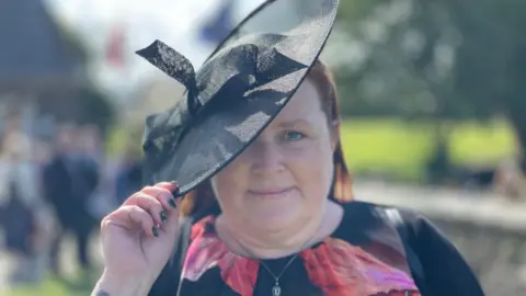 Kirstie Morphet, a woman a floral print dress, she wears a large black hat and tilts it as she smiles.