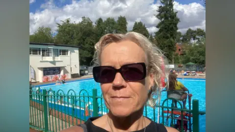 Deborah Aydon Aydon takes a selfie at Droitwich Spa Lido. She is wearing dark sunglasses and has tied back blonde hair. Behind her is a lifeguard in a yellow t-shirt watching over children playing in the water.