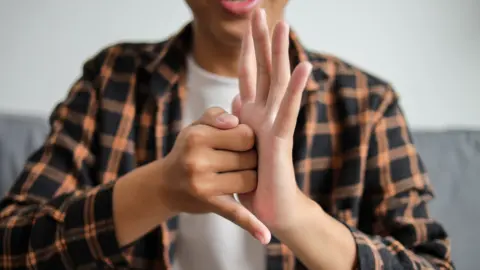 A person wearing a checked black and tan shirt using sign language. The left hand is pointed upwards and the right is against it with the little finger stuck out at the bottom.