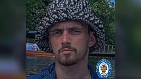 West Midlands polisi seorang pria dengan topi hitam dan putih berdiri di luar. Dia memiliki tunggul coklat dan kemeja biru.