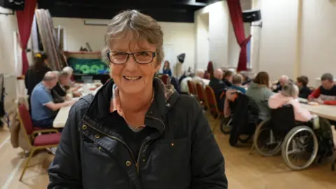 A woman with grey hair and glasses and a dark jacket on front of people sitting in a hall at tables opposite each other.