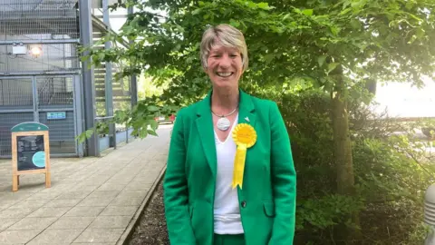 BBC/Emma Howgego Pippa Heylings looks at the camera with a smile on her face. She wears a green jacket, a yellow Liberal Democrat rosette and a necklace with a large circle pendant. In the background is a tree with lots of green leaves. To the left is a sign advertising coffee for sale.