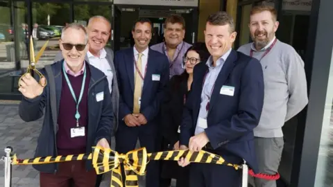 Gloucestershire College A group of people in front of a ceremonial ribbon, with the man on the left holding some golden scissors