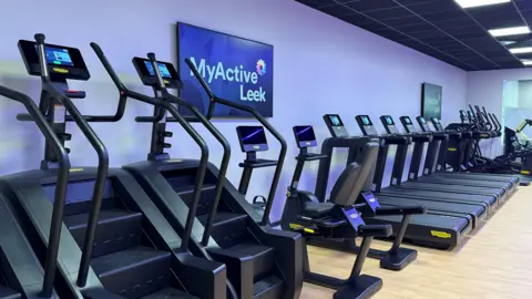 A gym with treadmills and cross trainers has a pale purple wall and a light wood-effect floor. There is branding on tv screens in the room, which has a dark ceiling and light fittings space evenly along its length.