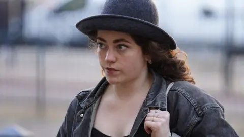 PA Media Emily Brocklebank with long brown hair wearing a dark bowler-type hat and a blue denim jacket, carrying a handbag