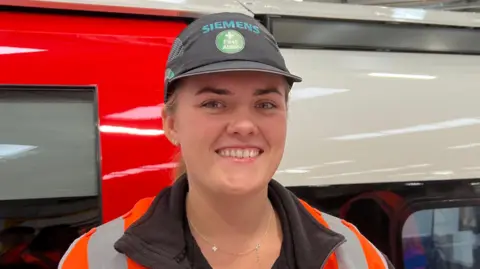 Pritti Mistry/BBC A woman is wearing a black Siemens' t-shirt with an orange hi-vis vest on and with a black Siemens' cap. She is smiling at the camera in front of a tube carriage that she is working on.