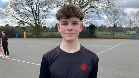 Oscar is stood in the school playground in a school branded black top with red piping and a red logo. He has mousy coloured, curly hair, and his hands behind his back.
