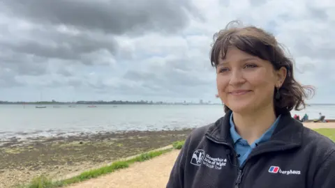 Image shows Kira looking off to the left of the picture. She is wearing a blue zip up fleece which says 'Hampshire and Isle of Wight Widlife Trust'. She is smiling. Her hair is brown and down to her shoulders. Behind her, is the water and you can see Portsmouth in the background. 