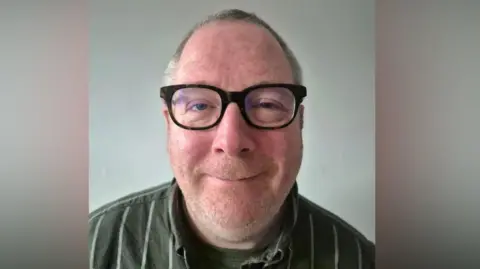 Supplied A man wearing glasses. He is also wearing a dark green top with light coloured stripes on it.