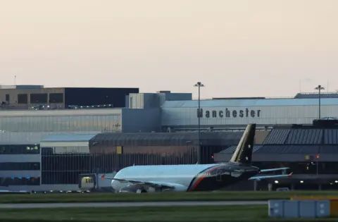 An airplane is on the runway, with grass in the foreground and an airport in the background with Manchester emblazoned on it