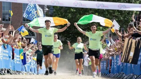 BBC/Phillip Stoneman Jordan and Cian Adams crossing the finish line wearing bright green tshirts, black shorts and running trainers. They are both waving green white and yellow Irish flags. Crowds stand behind metal barriers on both sides of a track. Two women in bright green t-shirts run behind them