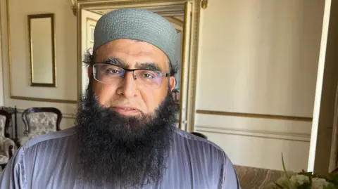 Mousumi Bakshi/BBC A man with glasses and a long black beard. He is wearing a hair covering and looking into the camera.