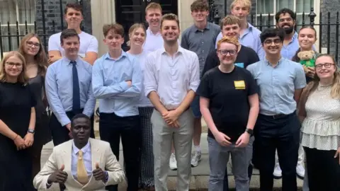 Un grupo de jóvenes posando para una foto frente a la entrada principal en Downing Street en Jack Abbott 10. Un hombre mayor se encuentra en el medio.