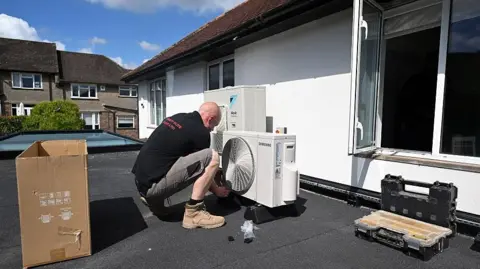 Getty Images Man installing a big air con unit on a roof of a house. There's an empty box and an open window on a white-coloured house.