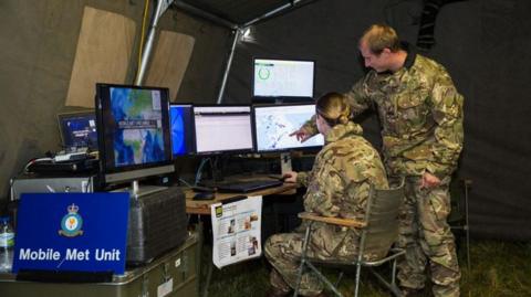 A Mobile Met Unit military tent with a desk and bank of computer screens. A female officer sitting at desk and a standing male officer pointing at a screen