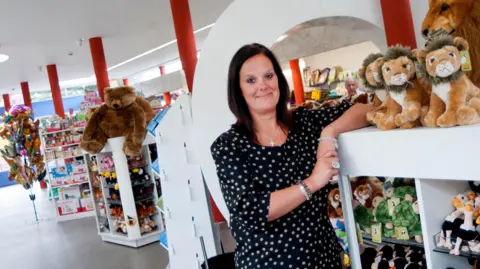 Dudley Zoo A woman with dark hair wearing a black and white polka dot top stands in a gift shop surrounded by shelves of soft toy animals. She is leaning against a white display unit with her arm resting on the counter. Stuffed lions, bears, turtles and other animals are visible on the shelves behind and beside her.
