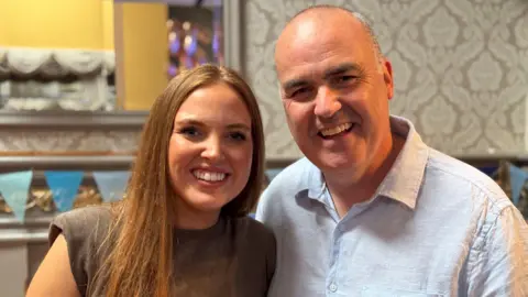 Molly and Andrew. Molly has long red hair, wearing a grey sleeveless top. Andrew is bald, wearing a white lined shirt.