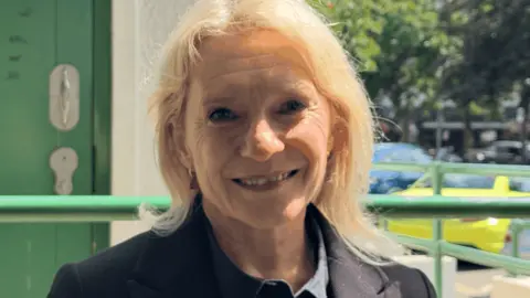 Senator Elaine Millar wearing a dark jacket, standing outside a public toilet entrance, with a green accessible restroom door visible in the background.
