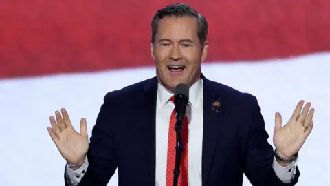 Reuters Rep. Mike Waltz stands in front of a red and white striped background wearing a blue blazer, white shirt and red tie. He is smiling and holding his hands up while speaking into a microphone