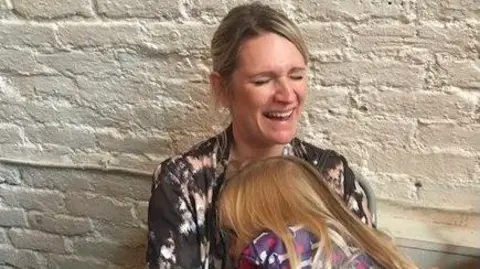 Geraint John Deb is laughing and holding her young daughter. Deb has fair hair which is died up and a patterned pink, white and black top. Her daughter has long blonde hair and has her head buried in her mother's chest. 