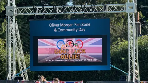 BBC A large screen has been put up on grey meal scaffolding. The screen is blue and reads "Oliver Morgan Fan Zone and Community Day." Below that writing is a photograph of Ollie Morgan within the Olympic rings, and it reads "Good Luck Ollie, we are with you every step of the way at Paris 2024"
