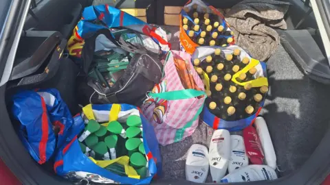 Policing Peterborough A large collection of bags in the back of a car, showing bottles of olive oil, toiletries, chocolate, and sprays. The boot of the car is open and items are placed on a grey piece of cloth. 