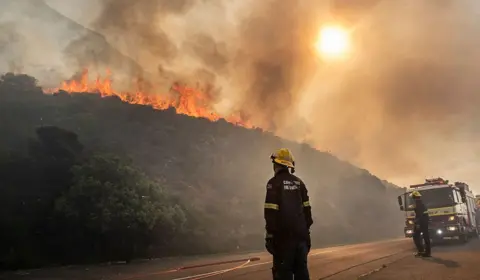 BRENTON GEACH / GALLO IMAGES/ GETTY IMAGES Firefighters look on as a fire blazes above the roadside. They have lorries and hose pipes which they are readying for use.

